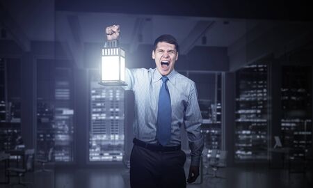 Screaming businessman holding glowing lantern on background office interior. Front view of emotional man in shirt and tie looking for something in dark. Scared business person walking with lanternの写真素材