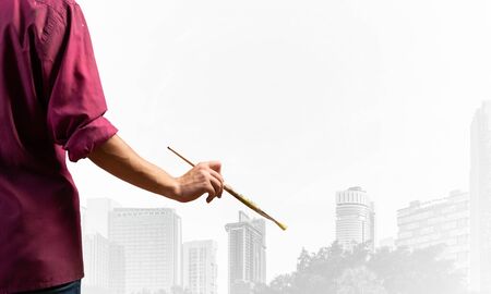 Close up artist hand holding paintbrush. Painter in shirt standing on background modern skyscrapers. Creative hobby and artistic occupation concept with copy space. Art imagination and inspirationの写真素材