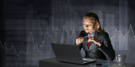 Charming businesswoman sitting at office desk and working at laptop on background grey wall. Smiling girl in business suit and tie busy at work in evening. Business accounting and investment analysisの写真素材