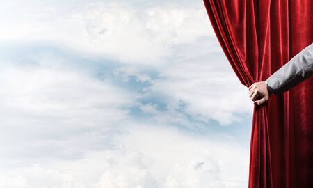 Human hand opens red velvet curtain on blue sky backgroundの写真素材