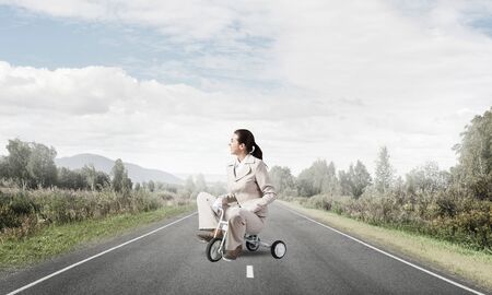Caucasian woman riding kids bicycle on asphalt road. Young employee in white business suit biking outdoor. metaphor of ineffective and incompetent work. Beginner level concept with bicyclist.の写真素材