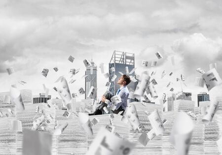 Thoughtful businessman looking away while sitting on pile of documents among flying papers with cityscape on background. Mixed media.の写真素材