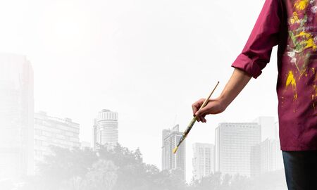 Close up artist hand holding paintbrush. Painter in shirt standing on background modern skyscrapers. Creative hobby and artistic occupation concept with copy space. Art imagination and inspirationの写真素材