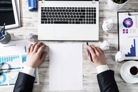 Top view modern office workspace with businessman hands and blank white paper. Flat lay white wooden desk with laptop keyboard, business charts and cup of coffee. Corporate business people at work.の写真素材