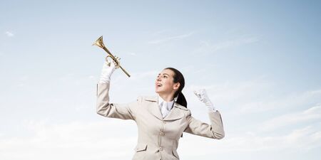 Beautiful woman holding trumpet brass overhead. Young smiling businesslady in white business suit and gloves posing with music instrument on blue sky background. Business concept with musicianの写真素材
