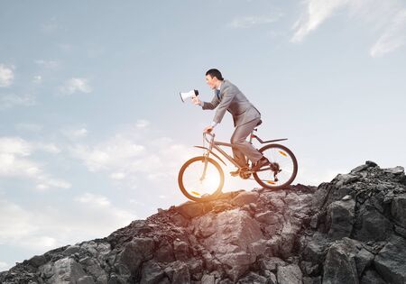 Businessman with megaphone in hand on bike at sunny day. Promotion and announcement. Corporate employee in business suit on bicycle riding downhill on mountain road. Cyclist on background of blue skyの写真素材