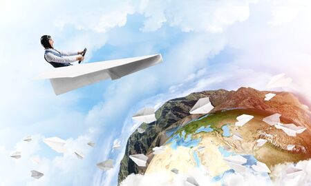 Pilot in leather helmet driving paper plane on background of blue cloudy sky. Funny man flying in small paper airplane. Spherical view of earth planet with lands, ocean and mountains.の写真素材