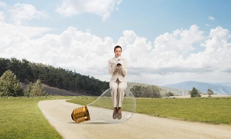 Attractive woman playing trumpet brass on asphalt road. Young businesslady in white business suit and gloves sitting on big light bulb with music instrument. Musician practicing and performing outdoorの写真素材