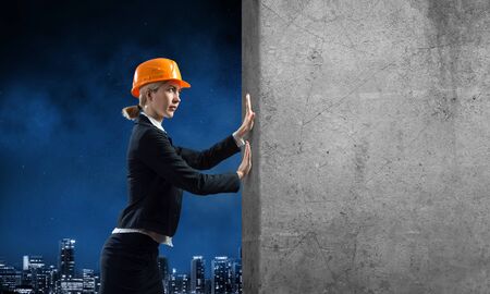 Beautiful woman architect pushing concrete wall. Young specialist in safety helmet on night city background. Overcoming obstacles and solution of problems. Grey concrete wall as banner with copy spaceの写真素材
