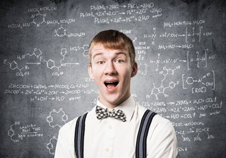 Happy teenager with broad smile. Emotional redhead boy doing mathematical calculations. Young scientist wears white shirt, bow tie and suspenders on background of chalkboard with math formulas.の写真素材