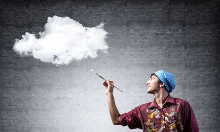 Young artist drawing realistic white cloud. Male painter in dirty shirt and bandana with paintbrush on grey wall background. Creative hobby and artistic occupation. Art classes concept with copy spaceの写真素材