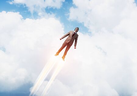Businessman in aviator hat flying in cloudy blue sky as superhero