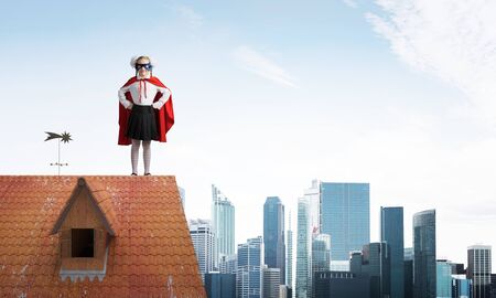 Young girl in superhero costume standing on house brick roof. Mixed mediaの写真素材