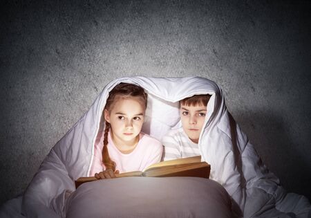 Little girl and boy looking at each other under blanket. Children reading magic stories in bed before going to sleep. Young sister and brother in pajamas together on background of grey wall.の写真素材