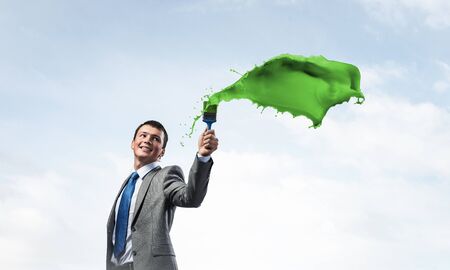 Smiling creative businessman painter holding paintbrush. Portrait of joyful handsome man in business suit and tie. Green paint splash on blue sky background. Ambitions and creativity in businessの写真素材