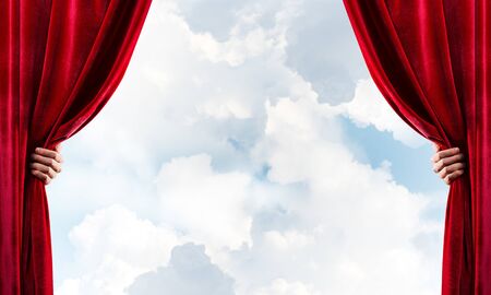 Human hand opens red velvet curtain on blue sky backgroundの写真素材