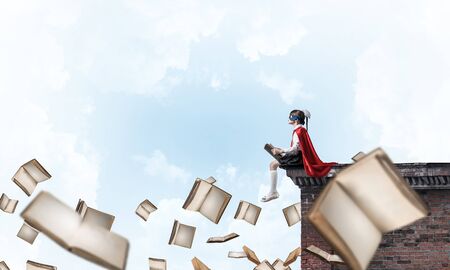 Little child in mask and cape sitting on building roof and reading . Mixed mediaの写真素材