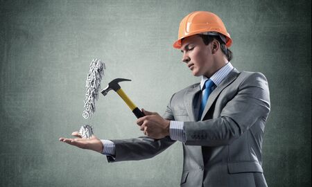 Businessman going to crash with hammer exclamation mark. Young handsome man in business suit and safety helmet standing on wall background. Search for solution and realization. Risk managementの写真素材