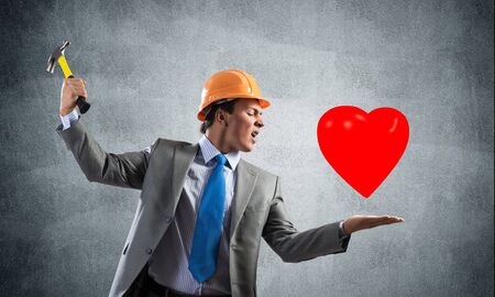 Furious businessman going to crash red heart with hammer. Young handsome man in business suit and safety helmet standing on grey wall background. Love treason and crisis in family relationships.の写真素材