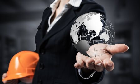 Businesswoman holds in palm 3d world globe. Woman in business suit with orange safety helmet. International company presentation of engineering and project management. Global financial system.の写真素材
