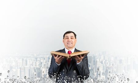 Happy businessman holding open old book. Smiling man in suit standing on city background with flying...