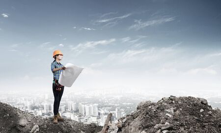 Female engineer in hardhat standing with technical blueprint on construction site. Young woman architect in workwear inspecting construction on background of cityscape. Building project management.の写真素材