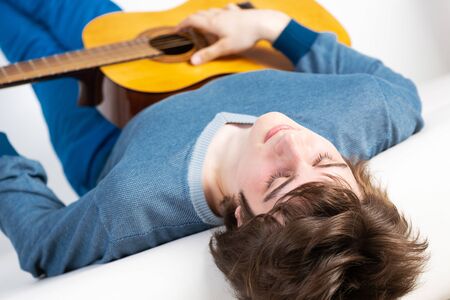Playing guitar as hobby concept. Happy young music player lying back on sofa with guitar. Smiling guitarist relaxing on white sofa at home. Close-up musician practicing acoustic guitar.の写真素材