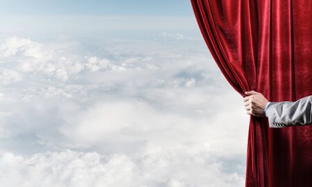 Human hand opens red velvet curtain on blue sky backgroundの写真素材