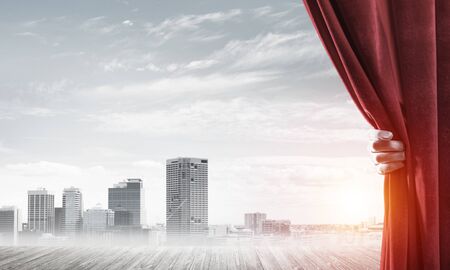 Human hand opens red velvet curtain to modern cityscape and cloudy skyの写真素材