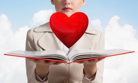 Beautiful woman showing red heart above opened notebook. Love and tenderness, valentines holiday and fall in love concept. Elegant girl in white business suit with open book on background of skyscape.の写真素材