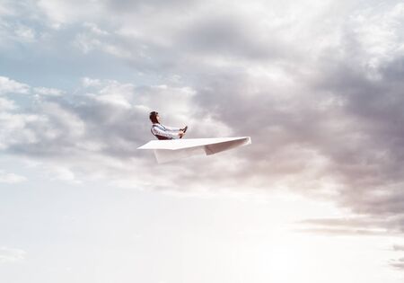 Aviator having fun in aircraft. Pilot in leather helmet and goggles driving paper plane in cloudy blue sky. Funny man flying in small paper airplane. Cloudscape background with fluffy clouds.の写真素材