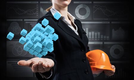 Businesswoman holds in palm abstract 3d cube model. Woman engineer in business suit with orange safety helmet. Engineering and project management. Architecture and construction company presentationの写真素材