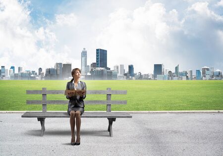 Young woman holding open book and sitting on wooden bench outdoors. Beautiful girl in business suit looking up on background modern cityline panorama in sunny day. Business education and growth.の写真素材