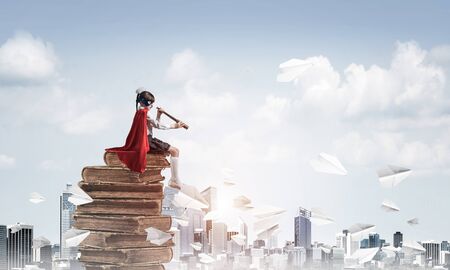 Little child in mask and cape floating on pile of books and looking in spyglassの写真素材