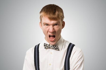 Frenzied hipster screaming with anger. Emotional redhead boy has furious facial expression. Portrait of guy wears white shirt, bow tie and suspenders on grey background. Emotion and expression conceptの写真素材