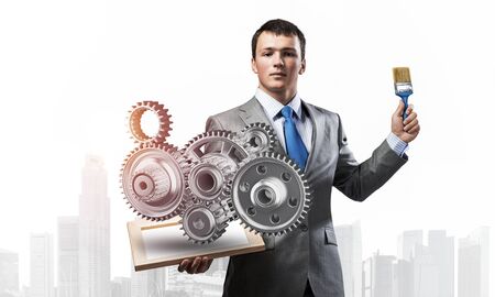 Creative businessman painter holding paint brush and 3d gears mechanism. Portrait of young man in business suit on city background. Presentation of mechanical engineering and project management.の写真素材