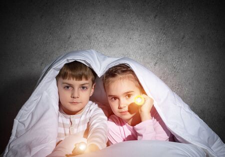 Children with flashlights lying in bed and looking in camera. Little sister and brother hiding under blanket together. Covered kids afraid of dark at night on background of grey wall.の写真素材