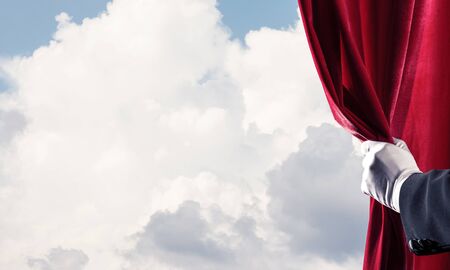 Human hand in glove opens red velvet curtain on blue sky backgroundの写真素材