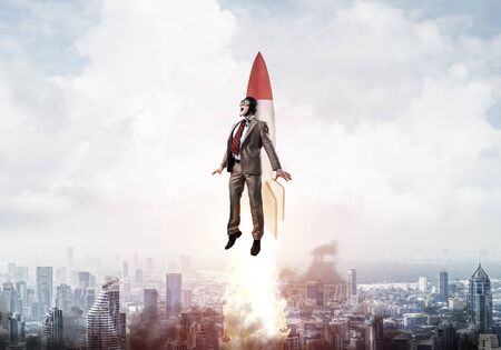 Businessman in suit and aviator hat flying on rocket. Superhero businessman flying with jetpack rocket in blue sky above modern downtown. Successful business startup. Career growth concept.の写真素材