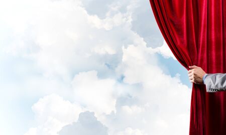 Human hand opens red velvet curtain on blue sky backgroundの写真素材