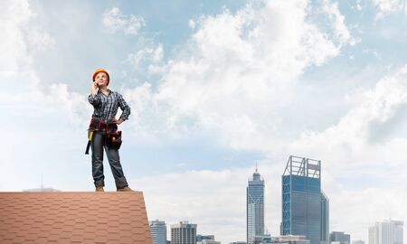 Attractive blonde woman in workwear and hardhat talking on smartphone. Young worker in checkered blue shirt standing on brick roof of building. Professional renovation and roofing construction.の写真素材