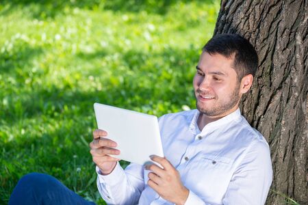 Man using tablet computer under tree in park on sunny day. Handsome guy looking at tablet screen. Happy smiling man sitting on green grass leaning against tree. Digital technology and communicationの写真素材