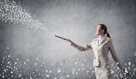 Woman looking at flying out random letters from opened book. Teaching and education. Information flow concept. Elegant young woman in white business suit with open notebook on background of grey wall.の写真素材