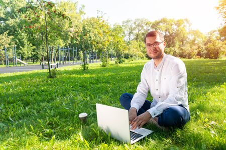 Young businessman using laptop computer on green grass in park. Handsome man in casual wear working with computer outdoors at sunny summer day. Freelance, blogging or remote work concept.の写真素材