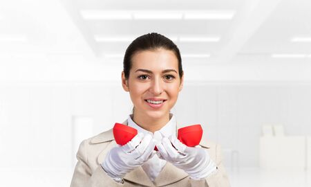 Smiling young woman holding retro red phone. Call center operator in white business suit posing with telephone in light office interior. Hotline telemarketing. Business assistance and consultation.の写真素材
