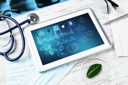 Laboratory patient examination in doctors office. Tablet computer with medical app interface on screen. Stethoscope, x-ray image and cardiogram on wooden desk. Medical diagnostics and examinationの写真素材