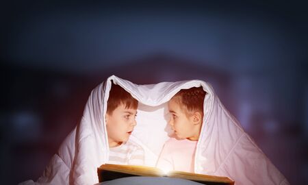 Scared little girl and boy looking at each other under blanket. Children together reading magic stories in bed before going to sleep. Young sister and brother in pajamas on deep blue sky backgroundの写真素材