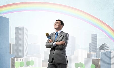 Confident and creative businessman painter standing with arms folded. Happy handsome man in business suit and tie holding paintbrush on city background. Cityscape illustration with colorful rainbow.の写真素材