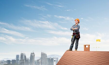 Female construction worker in hardhat standing with folded arms. Confident young woman in workwear standing on brick roof of building. Professional perspective and motivation. High-altitude workの写真素材
