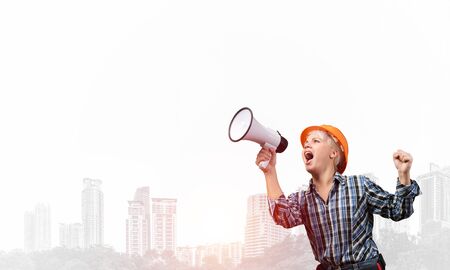 Expressive woman in safety helmet shouting into megaphone. Portrait of young emotional construction worker with wide open mouth on foggy city background. News announcement and advertisement.の写真素材